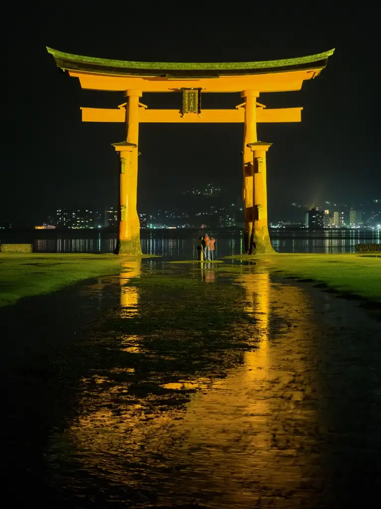 Three people look up in awe at the great sight of The O Torii Gate and the reflection of it with the tide out at midnight Miyajima Islanda