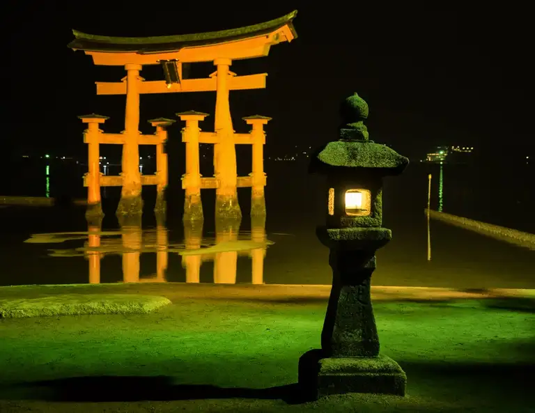 The O Torii Gate and stone lantern lit up in shadows of green and orange at midnight Miyajima Island