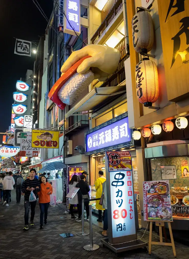 Giant fingers hold sushi over a couple walking through the streets of Dotombori Osaka Night time